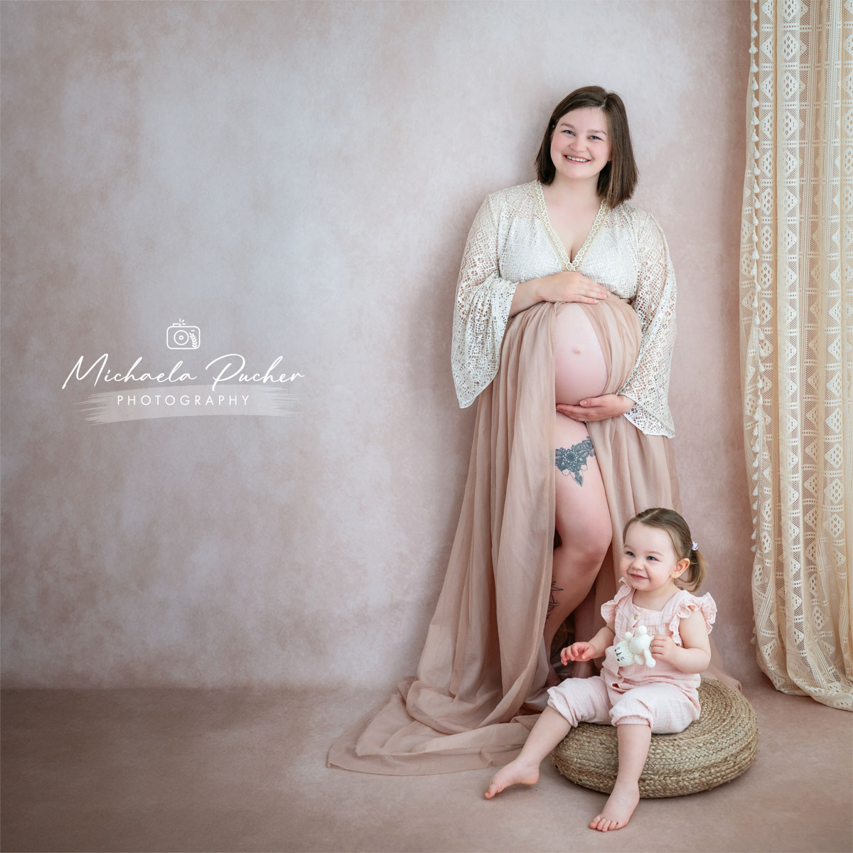 Hochschwangere Frau im Studio steht lächelnd vor einem hellen Hintergrund, hält ihren Babybauch und trägt ein langes, fließendes Tuch, während ihr kleines Kind davor auf einem runden Sitzkissen sitzt und fröhlich in die Kamera schaut.